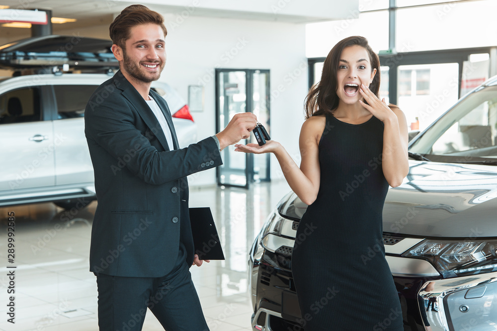 handsome man manager giving car keys to beautiful brunette woman client standing near brand new car after succesful deal in dealership center feeling happy handsome man manager giving car keys to beautiful brunette woman client standing near brand new car after succesful deal in dealership center feeling happy