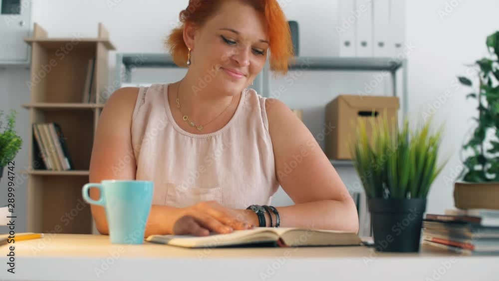 Young redhead woman in office read book with cup of tea or coffee. Relaxing on work with reading interesting books. 4k. Young redhead woman in office read book with cup of tea or coffee. Relaxing on work with reading interesting books. 4k.