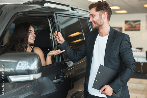 handsome man manager gives car keys to beautiful brunette woman client after succesful retail deal in dealership center standing near new purchased car