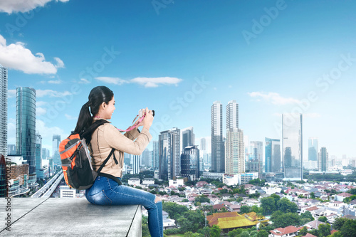 Asian woman with a backpack sitting on the rooftop and holding a camera to take pictures