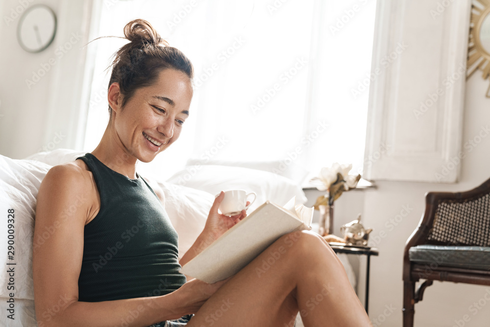 Pleased beautiful woman indoors at home sitting on floor near bed in a bedroom reading book. Pleased beautiful woman indoors at home sitting on floor near bed in a bedroom reading book.
