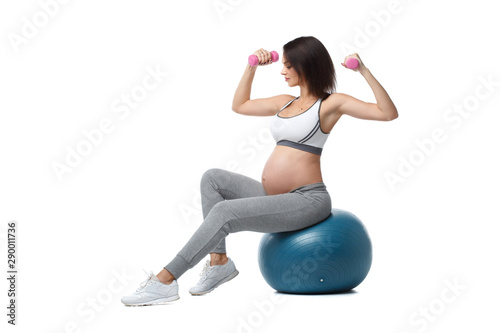 Slender athletic pregnant girl is engaged in fitness with dumbbells sitting on a ball isolated on a white background.