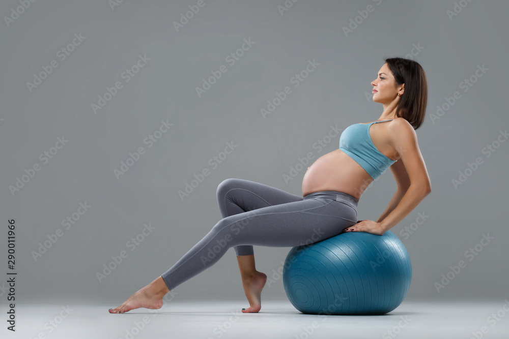 Slender athletic pregnant girl is engaged in fitness on a ball Isolated on a gray background.