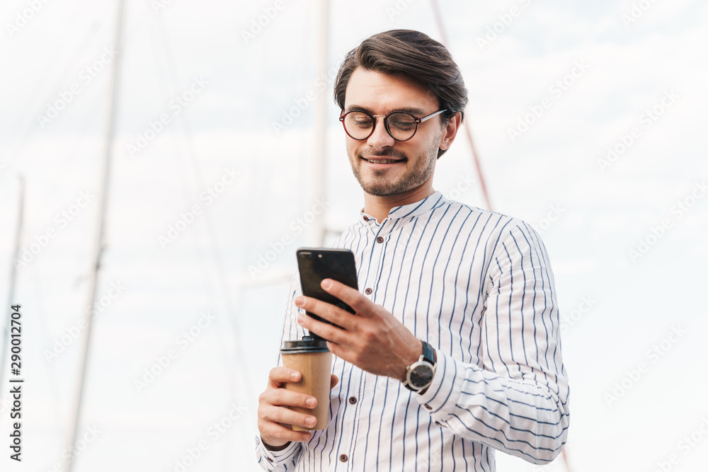 Photo of smiling young man typing on cellphone and drinking coffee from paper cup while walking on pier Photo of smiling young man typing on cellphone and drinking coffee from paper cup while walking on pier