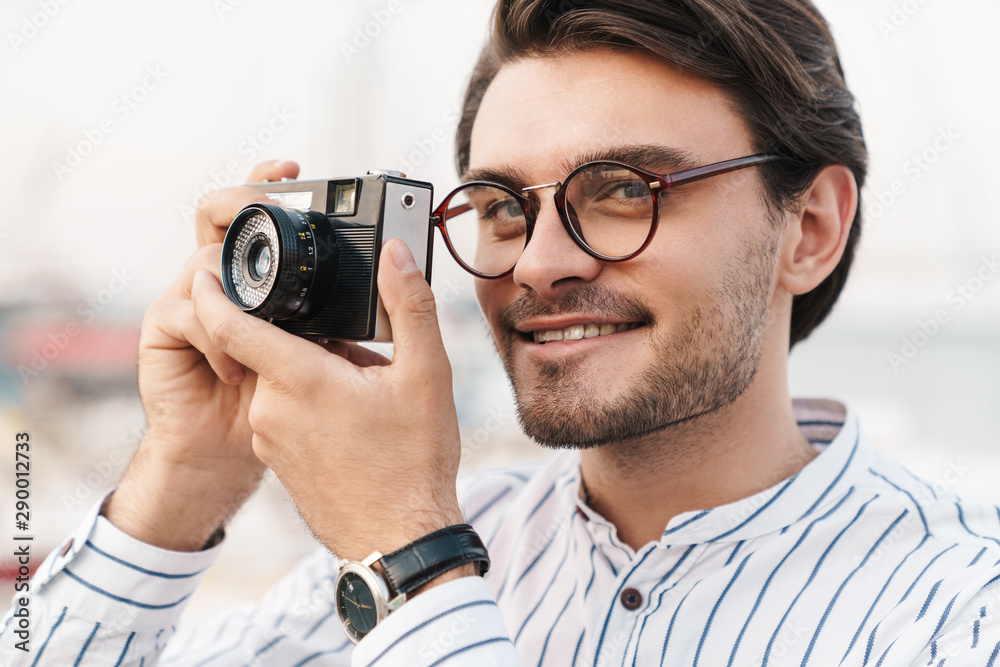 Photo of happy caucasian man taking photo on retro camera and smiling while walking on pier Photo of happy caucasian man taking photo on retro camera and smiling while walking on pier