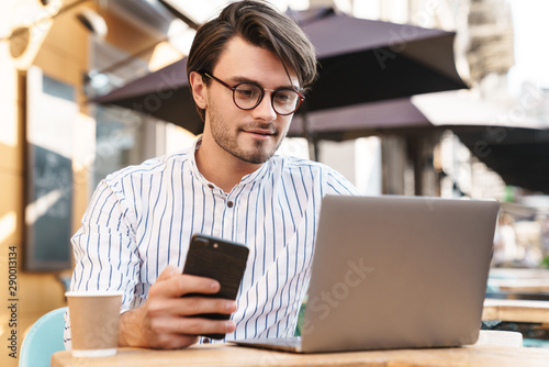 Photo of concentrated caucasian using laptop and cellphone while working in cafe outdoors