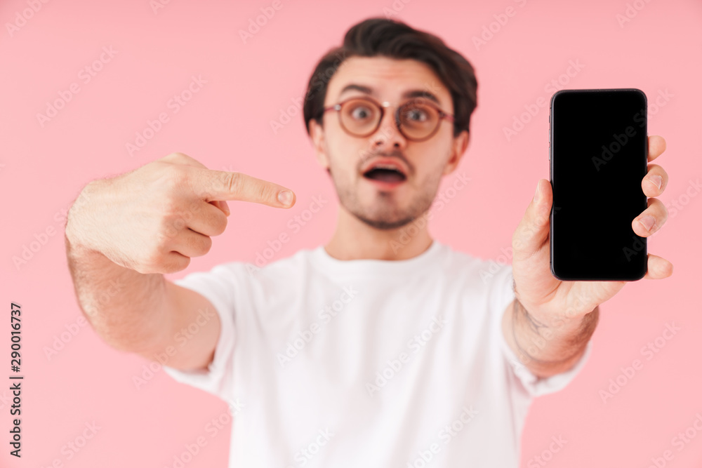 Image of young surprised man wearing eyeglasses holding and pointing finger at cellphone Image of young surprised man wearing eyeglasses holding and pointing finger at cellphone
