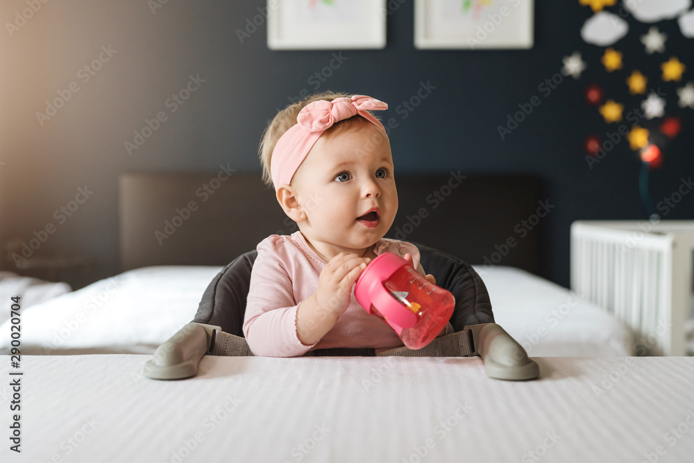Nine-month-old smiling baby girl in pink bandage sits at white table in highchair and drinks water from baby's cup. - obrazy, fototapety, plakaty 