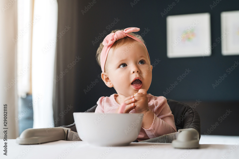 Nine-month-old smiling baby girl sits at white table in highchair and eats herself with spoon from bowl. Blurred background. Nine-month-old smiling baby girl sits at white table in highchair and eats herself with spoon from bowl. Blurred background.