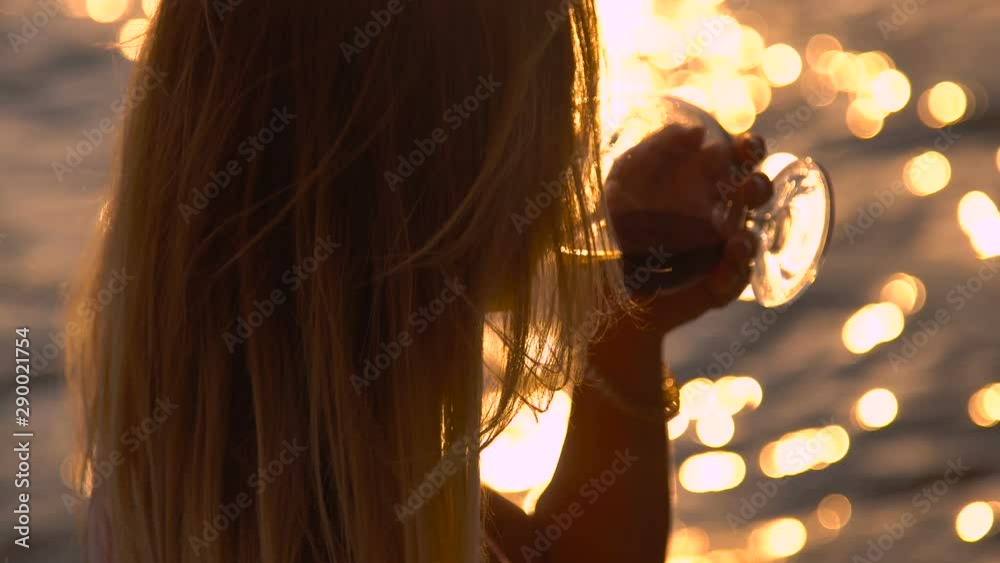 Young woman drinks red wine from a glass, sunset by the sea, close-up ...