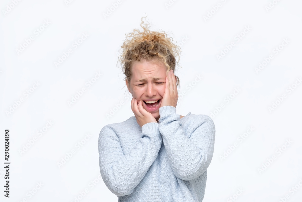 Frustrated young blonde curly woman in blue sweater having terrible headache keeping hands on head frowning her face with pain. Studio shot