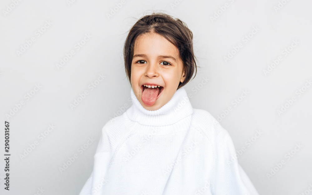 Funny kid little girl showing tongue, wearing big white knitted sweater, posing over studio light gray background on a holiday. Beautiful child has fun, during waiting for Christmas day. Funny kid little girl showing tongue, wearing big white knitted sweater, posing over studio light gray background on a holiday. Beautiful child has fun, during waiting for Christmas day.