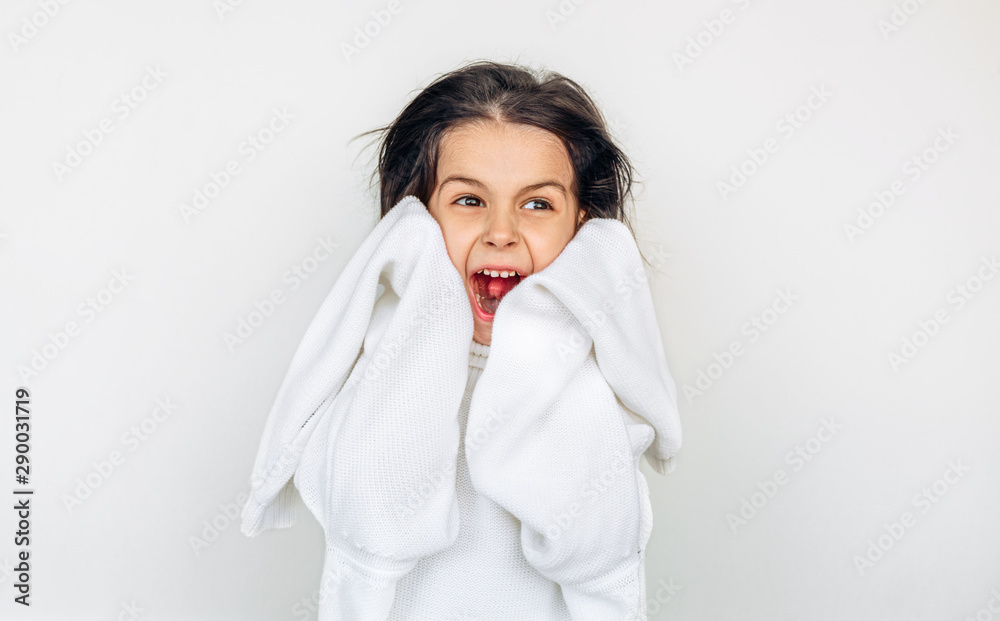 Amazed little girl has joyful expression and screams loudly, peeks out of big white knitted sweater, posing over studio light gray wall on a holiday. Happy child waiting for Christmas gifts. Amazed little girl has joyful expression and screams loudly, peeks out of big white knitted sweater, posing over studio light gray wall on a holiday. Happy child waiting for Christmas gifts.
