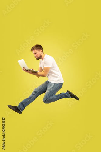 Full length portrait of happy jumping man isolated on yellow background. Caucasian male model in casual clothes. Freedom of choices, inspiration, human emotions concept. Using tablet in flight.