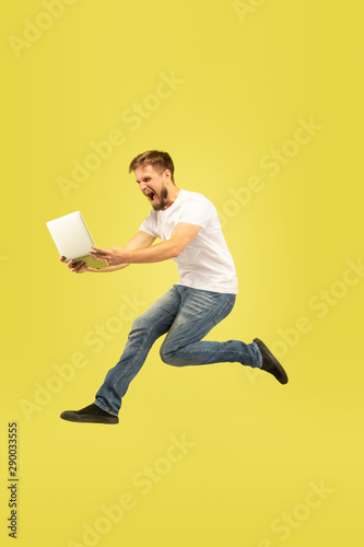 Full length portrait of happy jumping man isolated on yellow background. Caucasian male model in casual clothes. Freedom of choices, inspiration, human emotions concept. Using tablet in flight.