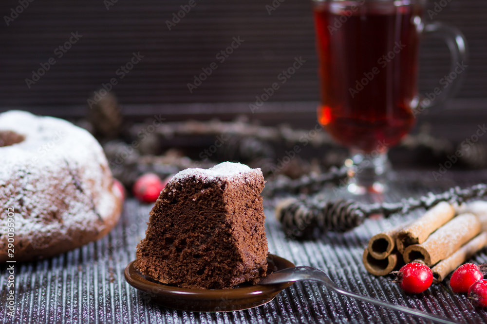 Piece of christmas chocolate sponge cake with sugar powder on wooden table with glass of red tea and sticks of cinnamon