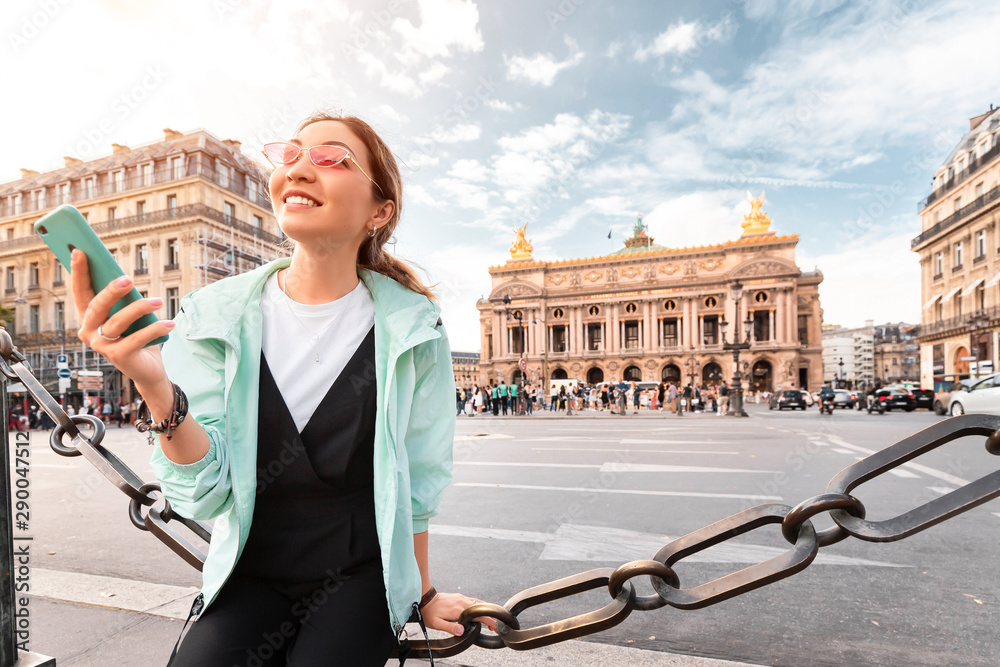 happy Asian girl traveler with phone near the main Facade of the Opera Garnier in the historic building of the Academy of music of Paris. The concept of Sights and cultural attractions of France - obrazy, fototapety, plakaty 