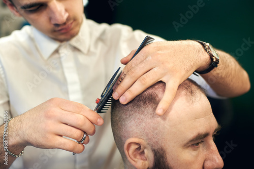 Cropped of male young hairdresser barber making short haircut for his client in modern barbershop. Concept of traditional haircutting with scissors. Step-by-step lesson for creating men's hairstyles.
