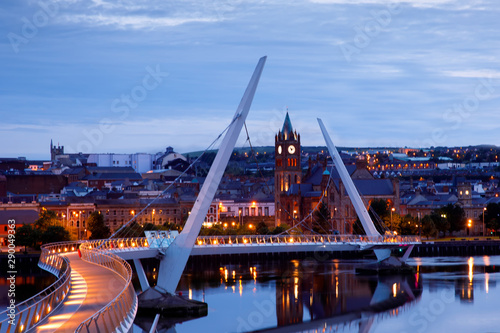 Derry, Ireland. Illuminated Peace bridge in Derry Londonderry, City of Culture, in Northern Ireland with city center at the background. Night cloudy sky with reflection in the river at the dusk photo