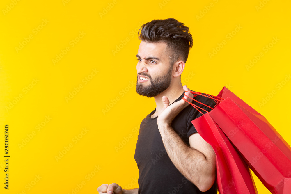 Side view of an upset young bearded stylish hipster man holding shopping bags posing on a yellow background. Concept of superfluous purchases. Side view of an upset young bearded stylish hipster man holding shopping bags posing on a yellow background. Concept of superfluous purchases.