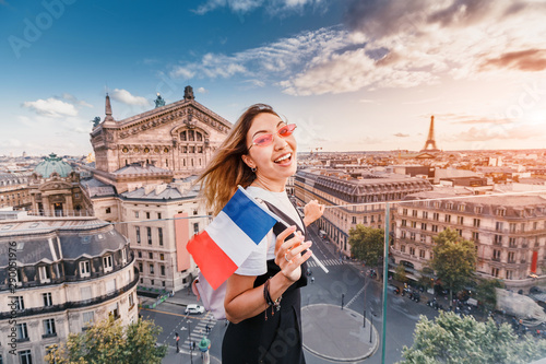 Fotografía Happy asian woman with french flag and backpack standing on a roof top and enjoys great view over Parisian skyline at sunset