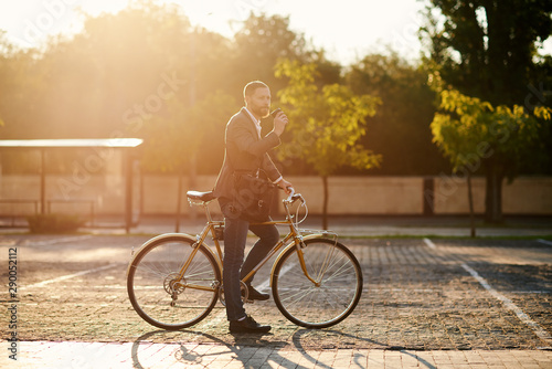 Portrait of handsome young red bearded man in casual clothes using phone, drink coffee and looking away and smiling while leaning on his bike, standing outdoors. Office and business center background