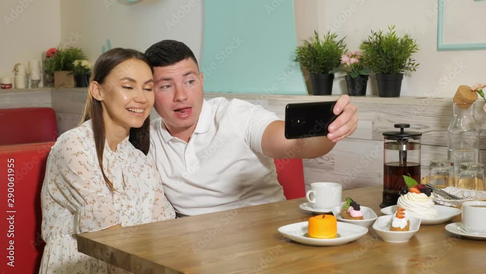 happy young couple girl guy makes selfie on modern smartphone sitting at served with cakes and drinks table in cafe happy young couple girl guy makes selfie on modern smartphone sitting at served with cakes and drinks table in cafe
