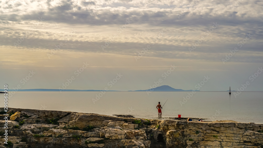 Obraz premium On an early cloudy morning, a fisherman catches fish from a rocky shore. The buoy, which is visible on the right, looks like a big float