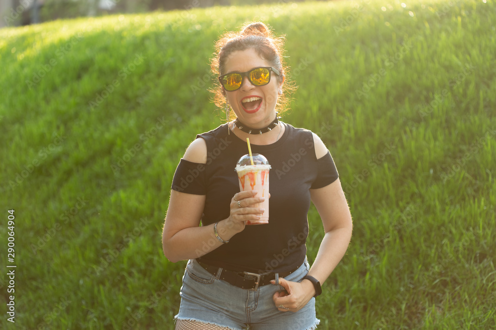 Fototapeta premium Portrait of a positive young pretty girl in punk clothes and glasses with a milkshake in her hands posing in a summer park on a warm summer evening. Vacation getaway concept.