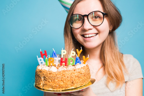 Close up funny positive girl in glasses and greeting paper hat holding a happy birthday cake in her hands standing on a blue background.