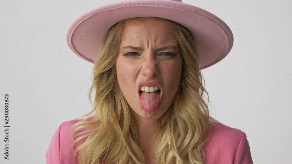 Close up view of unhappy young blonde woman in pink jacket and hat becoming disgusted and showing her tongue while looking at the camera over gray background isolated