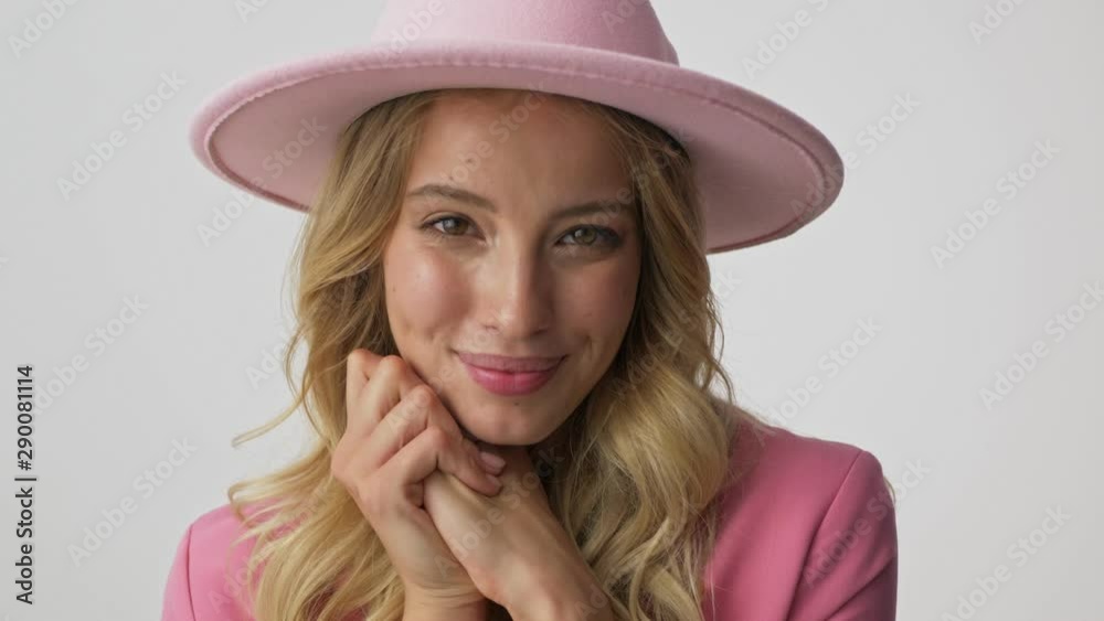 Close up view of smiling young blonde woman in pink jacket and hat blowing a kiss with two hands while looking at the camera over gray background isolated Close up view of smiling young blonde woman in pink jacket and hat blowing a kiss with two hands while looking at the camera over gray background isolated