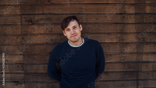 A handsome man in a blue sweater poses against a wooden background
