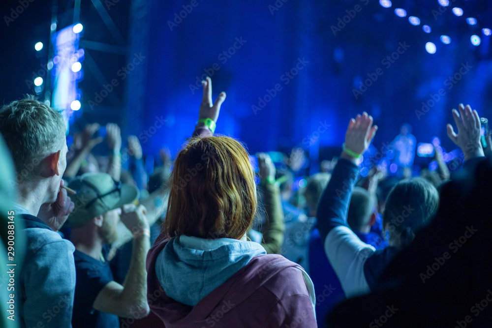 A crowd of young people at an evening street concert dancing and raising their hands up, illuminated by blue spotlights