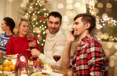 holidays, communication and celebration concept - happy young man calling on smartphone and having christmas dinner with friends at home