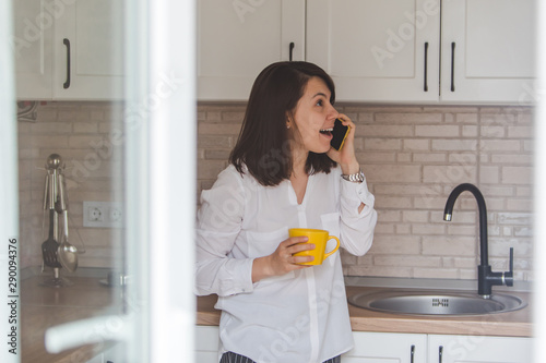 smiling happy woman talking no phone drinking tea from yellow mug at the kitchen