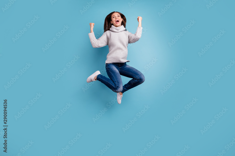 Full length photo of ecstatic content lady have holidays jump raise fists scream yeah celebrate victory wear white modern look pullover denim jeans sneakers isolated over blue color background