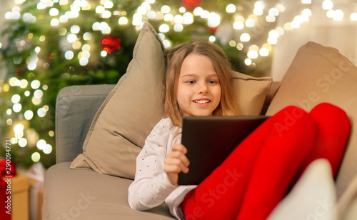 christmas, holidays and childhood concept - smiling girl with tablet pc computer lying on sofa at home