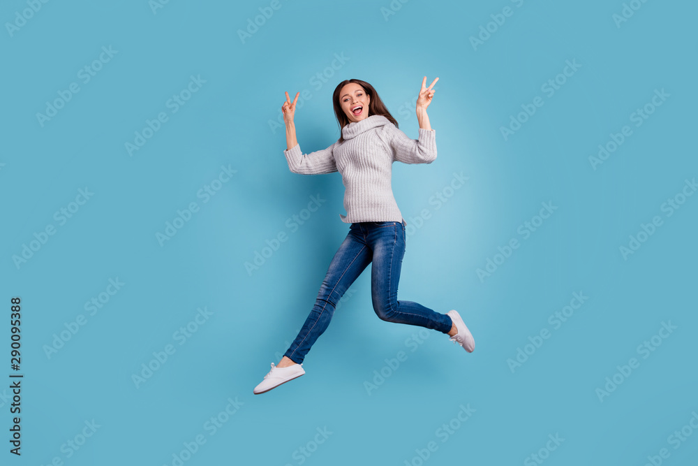 Full size photo of crazy content carefree girl jump make v-signs scream feel excited rejoice wear knitted pullover sweater good look denim jeans sneakers isolated over blue color background