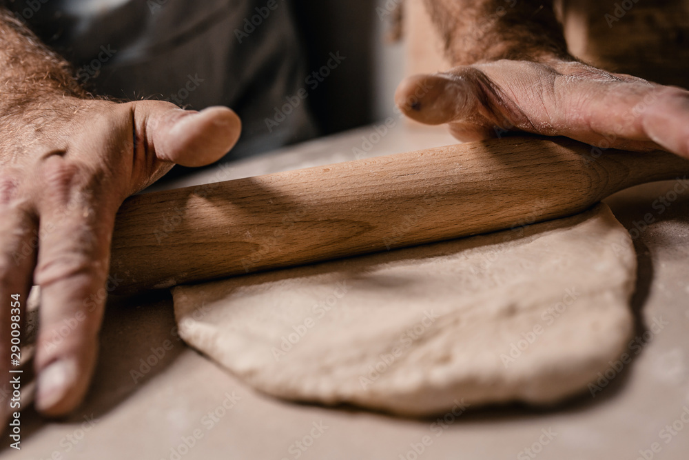 A baker hands with a wooden rolling pin rolls the dough into a thin layer on the table A baker hands with a wooden rolling pin rolls the dough into a thin layer on the table