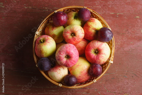 Early red apples and plums in a wooden basket, top view