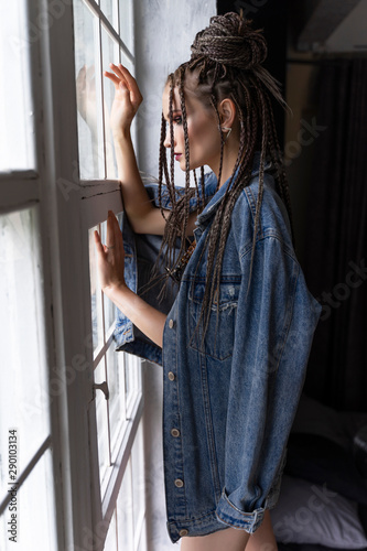 A beautiful young girl with an afro cornrows hairstyle, wearing a casual denim jacket and shorts, relaxes by the window and looks outside thoughtfully. Fashionable, commercial, modern design.