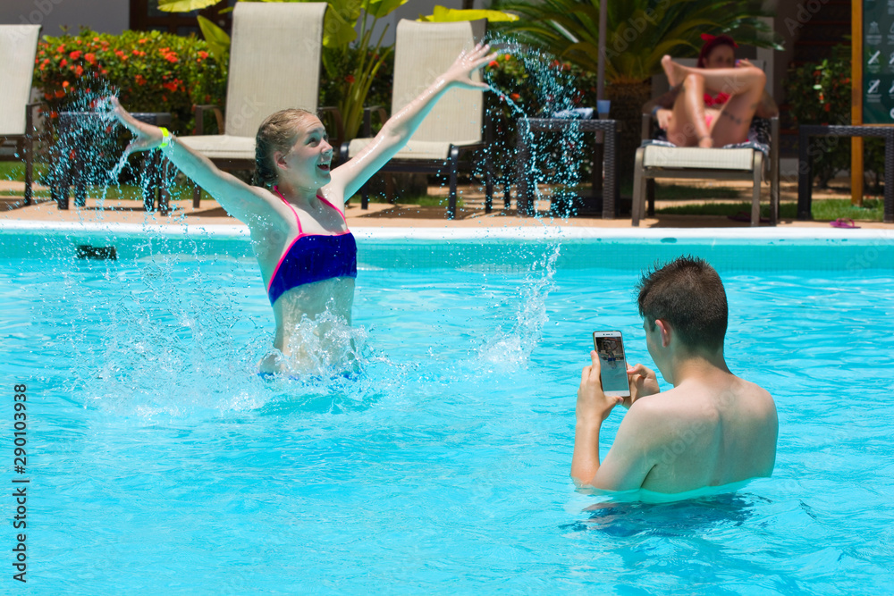 Young boy taking photos of teenage girl in luxury hotel swimming pool. Summer vacation concept