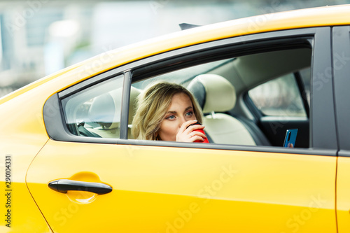 Photo of young woman drinking coffee while sitting in back seat of yellow taxi in summer on modern city background