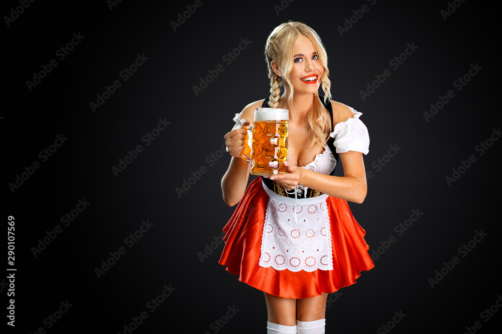 Smiling young sexy oktoberfest girl waitress, wearing a traditional Bavarian or german dirndl, serving big beer mug with drink isolated on black background. - obrazy, fototapety, plakaty 