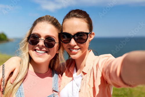 leisure and friendship concept - happy smiling teenage girls or best friends in sunglasses hugging and taking selfie at seaside in summer