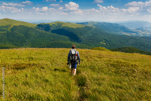 mężczyzna z plecakiem wędrujący po górach zielonych porośniętych trawą, widziany od tyłu