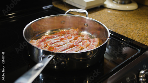 Bacon frying in stove top pan. Sizzling bacon strips in silver skillet.