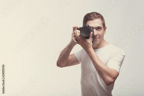 Smiling Man with camera isolated on light background. Young man holding digital camera and making photo looking on you. Lifestyle, travel and technology concept. Happy boy in white t-shirt with dslr