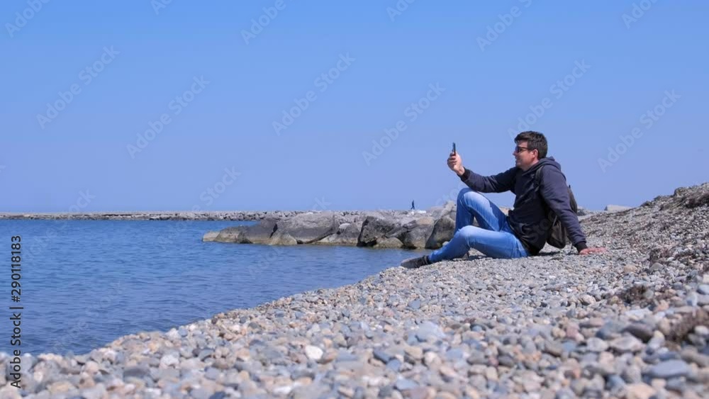 Man talks video chat in his smartphone sits on sea stone beach on vacation. Guy tourist on vacation. He is with backpack wearing jeans and sweatshirt in sunglasses. Traveller call mobile phone.